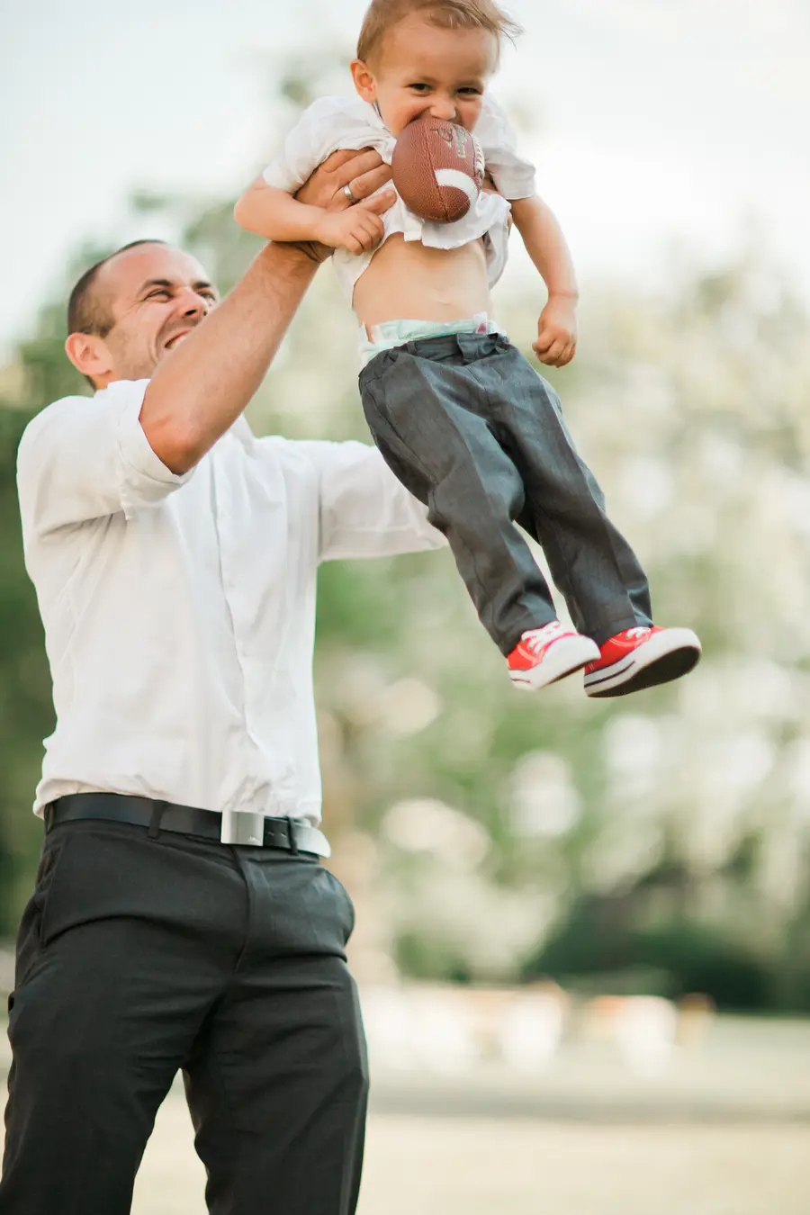 A man in a white dress shirt and black pants throws a young child, wearing a white shirt and gray pants, into the air. The child has a small football in their mouth.