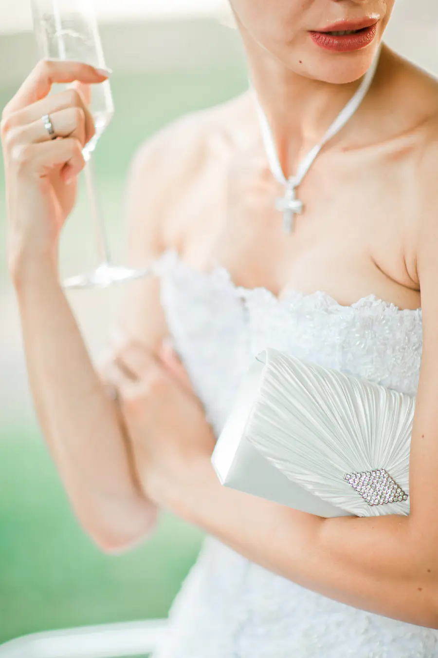 A bride in a white strapless dress holds a champagne flute in one hand and a white clutch purse adorned with rhinestones in the other.