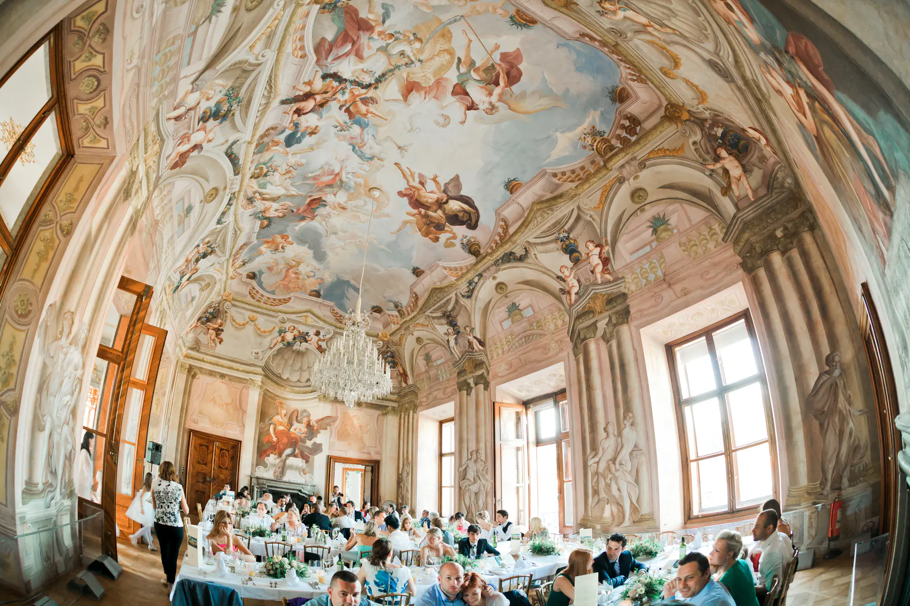 A wide-angle view of a grand, ornately decorated ballroom with a frescoed ceiling depicting cherubs and mythological figures. Guests are seated at long, white-clothed tables, enjoying a meal.