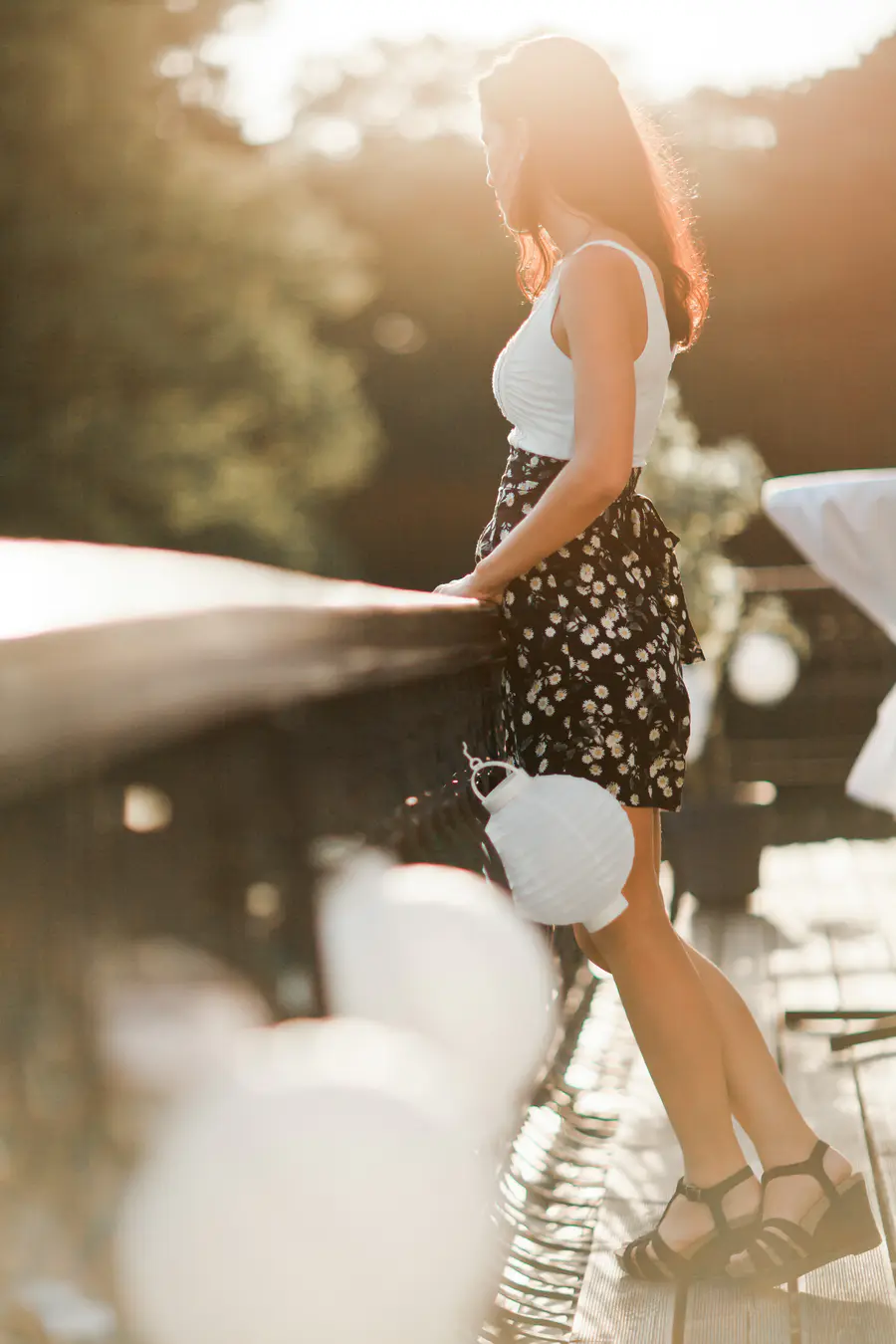 A woman in a white tank top and a floral skirt stands with her hands on a wooden railing, looking out at a sun-drenched landscape.