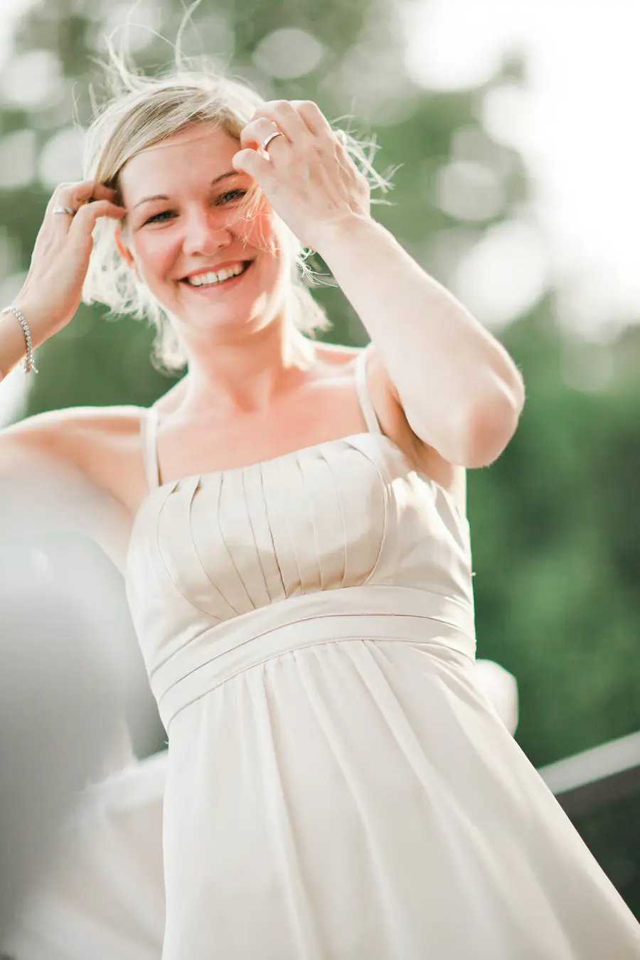 A blonde woman smiles as she touches her hair with both hands, wearing a light-colored, strappy dress.