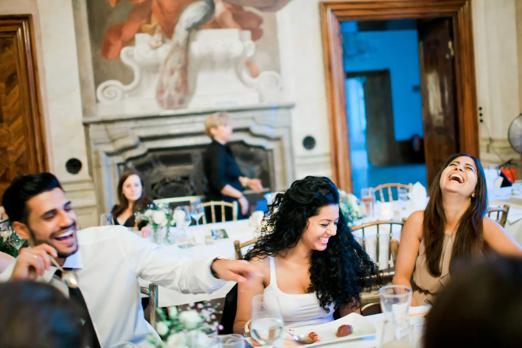 A group of people are laughing heartily around a dinner table in a formal setting, with a woman in the foreground laughing the loudest.