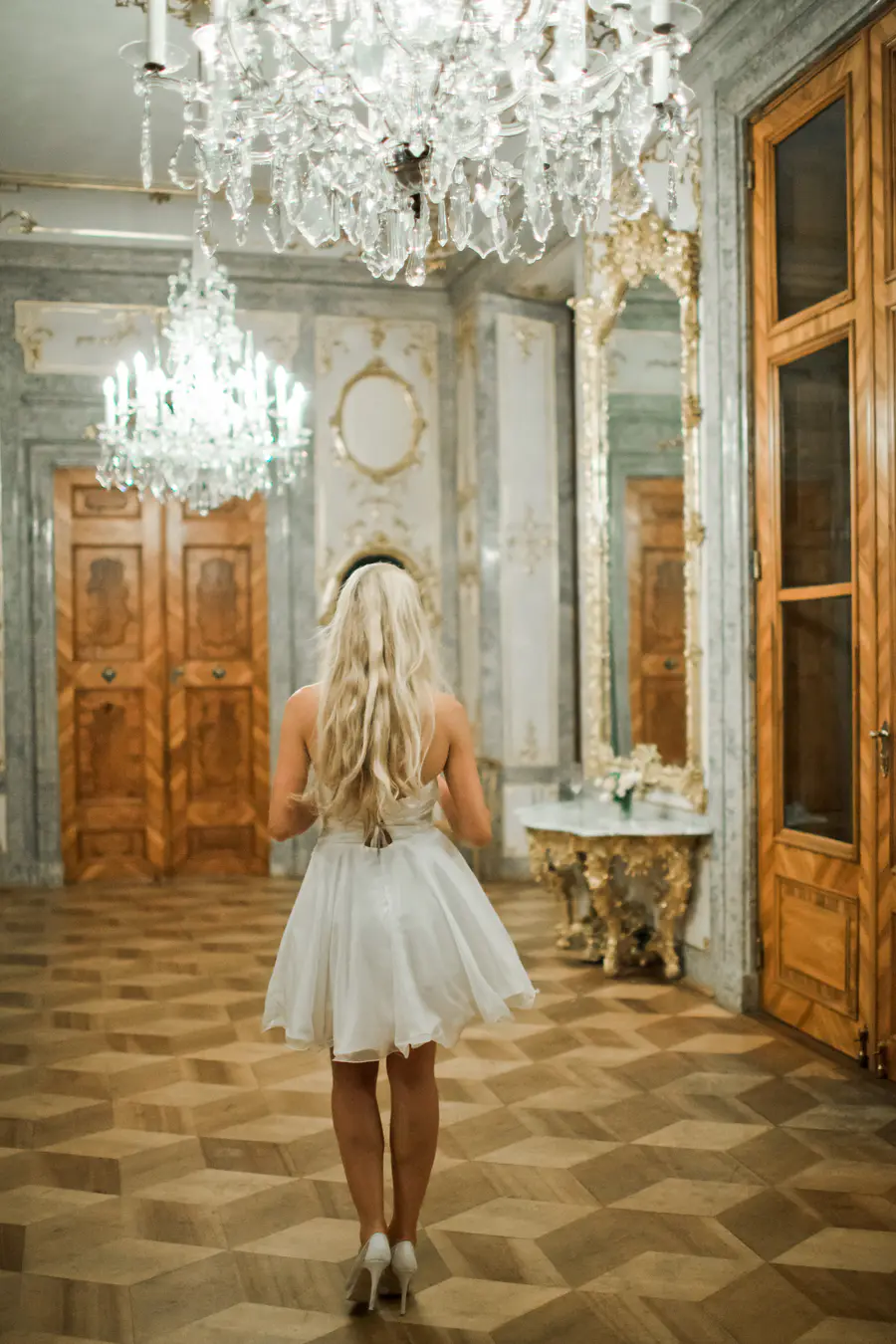 A woman with long blonde hair, wearing a short white dress and white high-heeled shoes, walks away from the camera in a grand, ornate ballroom.
