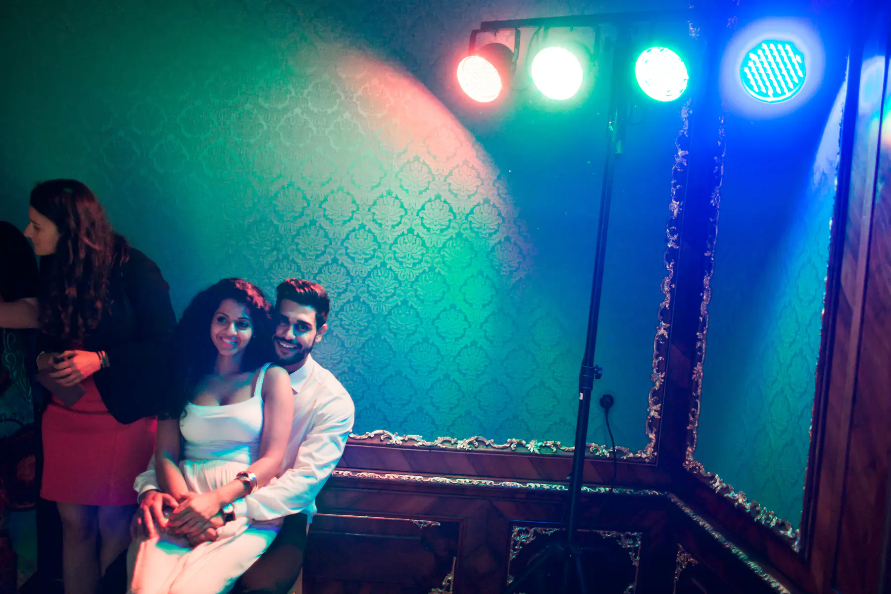A man and woman embrace in front of a patterned teal wall illuminated by colorful stage lights.