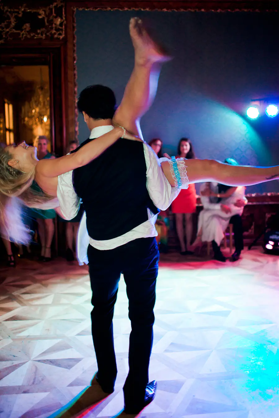 A man in a white shirt and black vest dips a laughing blonde woman, holding her legs up in the air, as they dance on a patterned floor lit by colorful lights.