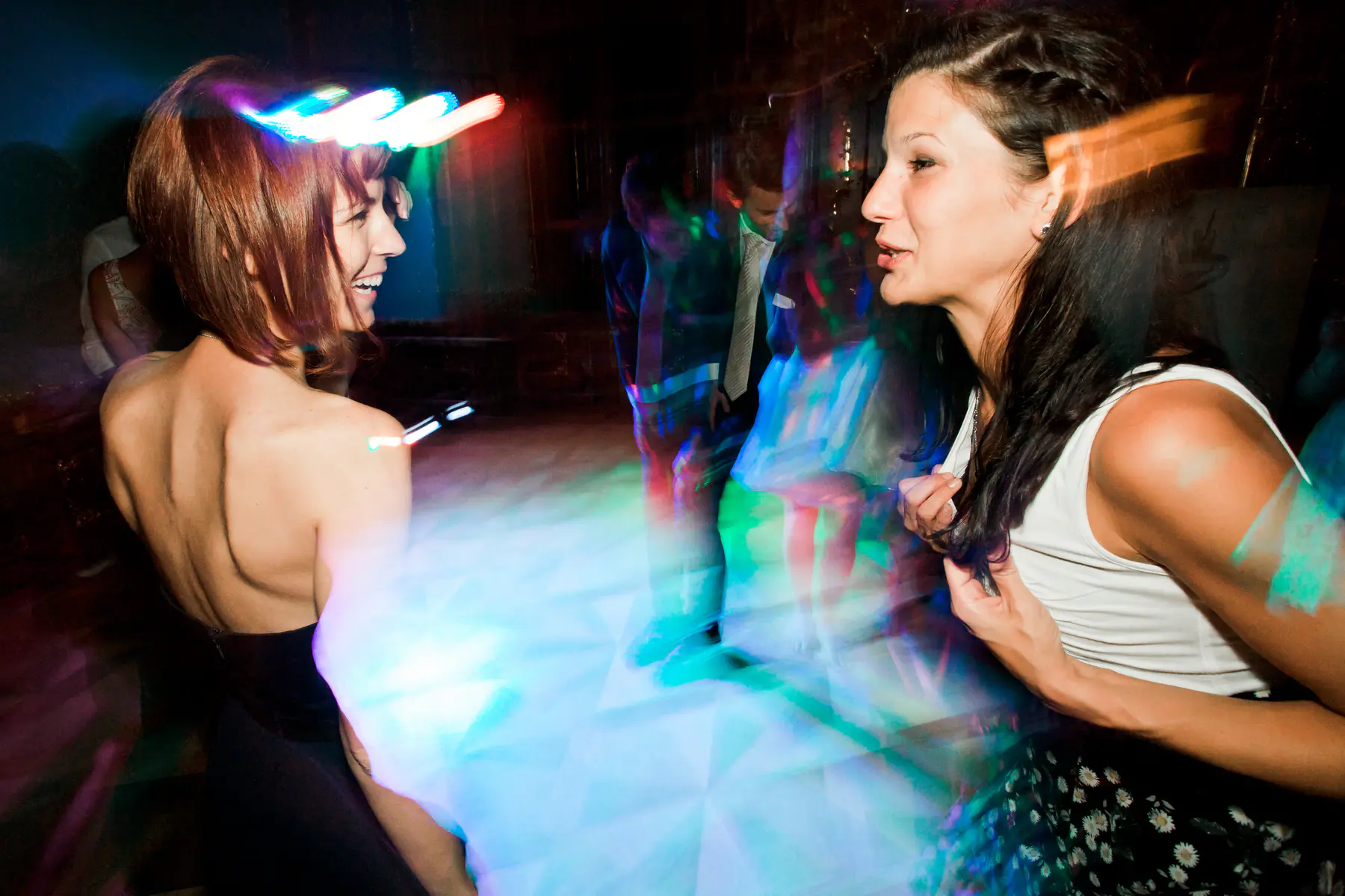 Two women with blurred faces are talking and laughing at a party with colorful lights.