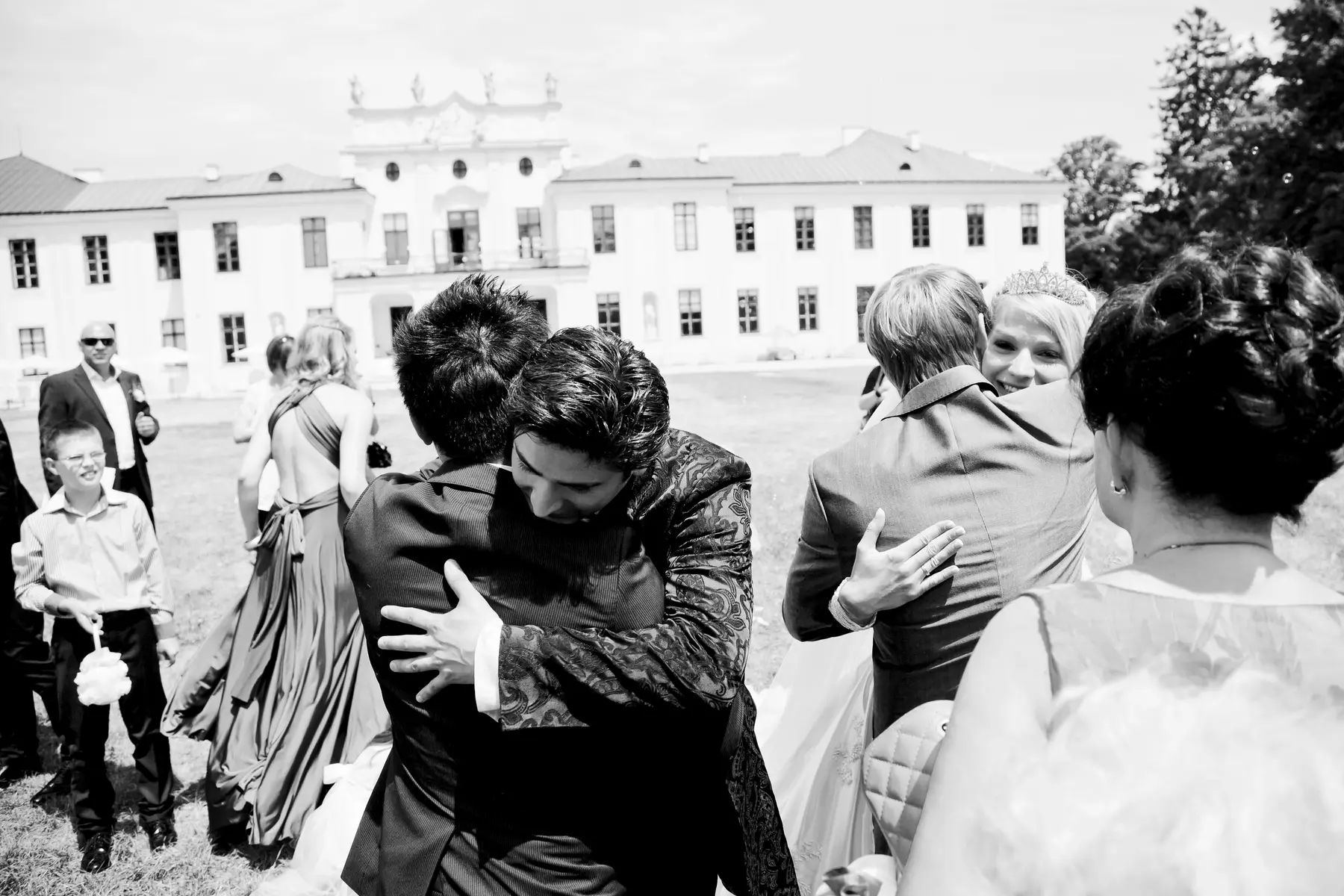A group of people in formal attire hug outside a white building, likely at a wedding.