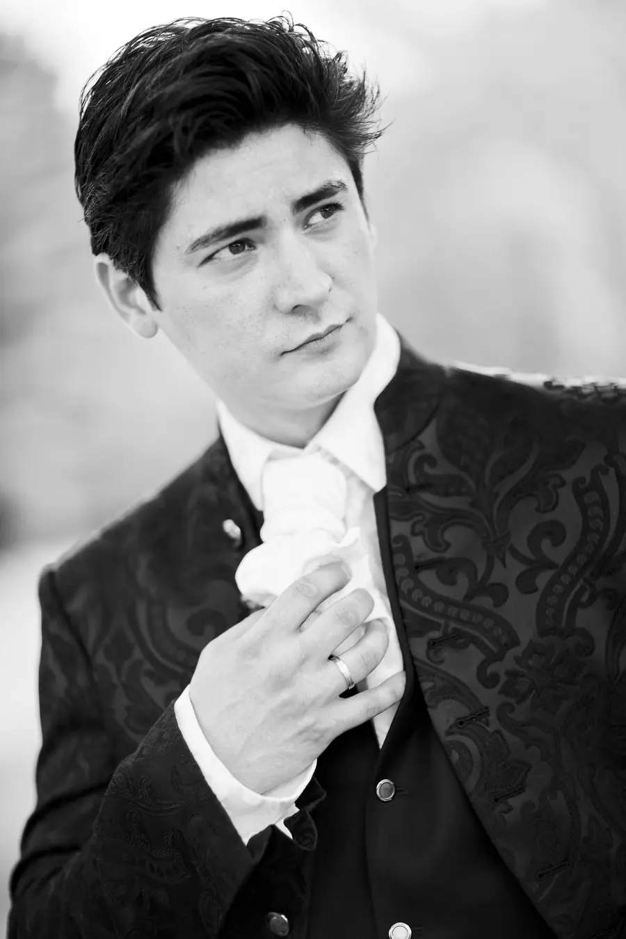 A black and white, waist-up portrait of a young man in a formal suit. He has dark, styled hair and is looking off to the right with a slight smile. He's wearing a white ruffled shirt, a dark vest with silver buttons, and a dark jacket with an ornate pattern. His right hand is holding the ruffled shirt near his chest, and he wears a ring on his left ring finger. The background is blurred and light.