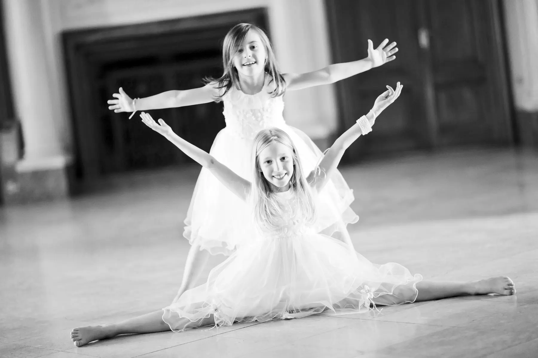 Two young girls in white dresses pose with arms outstretched in a black and white photograph. The girl in front is doing a split on the floor, and the girl behind her is standing.