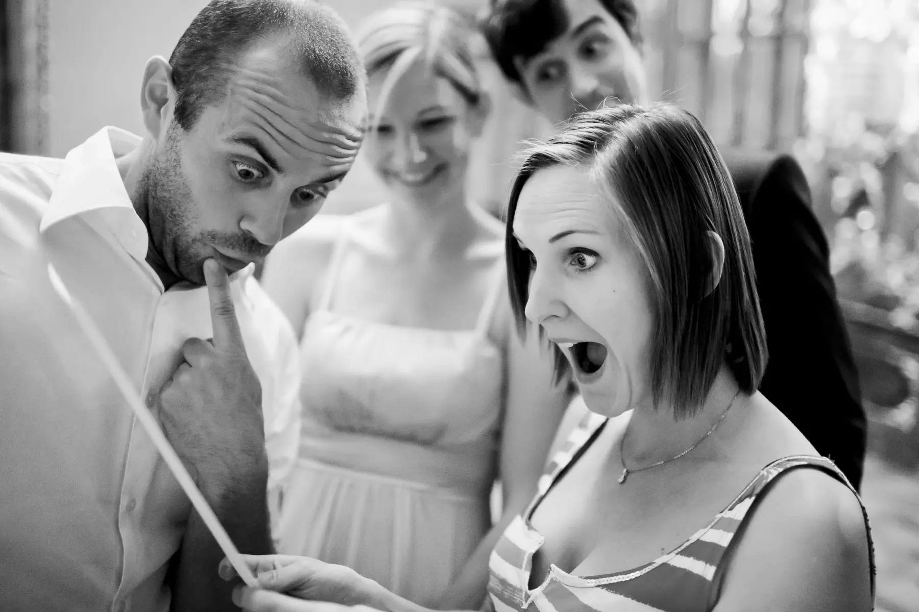 Four people, two men and two women, are looking intently at wedding menu held by the woman on the right. The woman on the right has her mouth open in surprise and her eyes wide. A man on the left is looking down and to the right with a surprised expression, his finger to his lips. A woman in the background is smiling, and a man behind her has a wide-eyed, surprised expression.