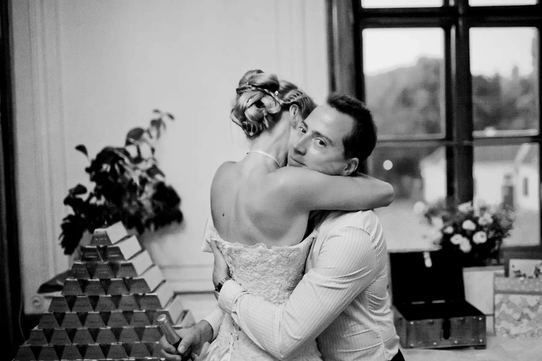 A friend hugs bride from behind while she holds a champagne bottle, with a pyramid of gold bars in the foreground and a window in the background.