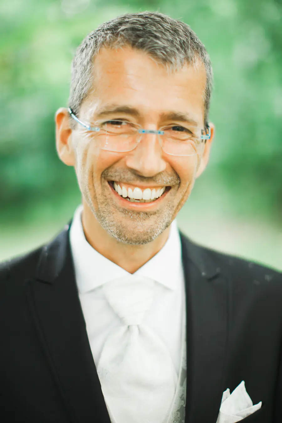 A man in a black suit and white tie smiles warmly at the camera. He is wearing clear-rimmed glasses with blue accents. The background is blurred green foliage.