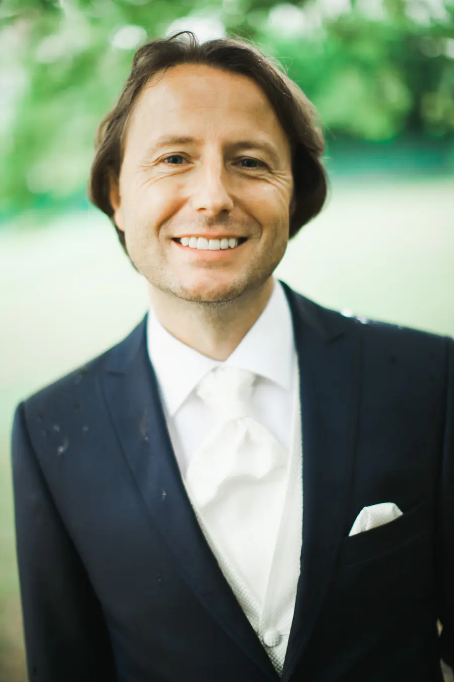 A man in a black suit and tie smiles at the camera, with a white rose boutonnière pinned to his lapel. He stands outdoors with soft sunlight and trees in the background.