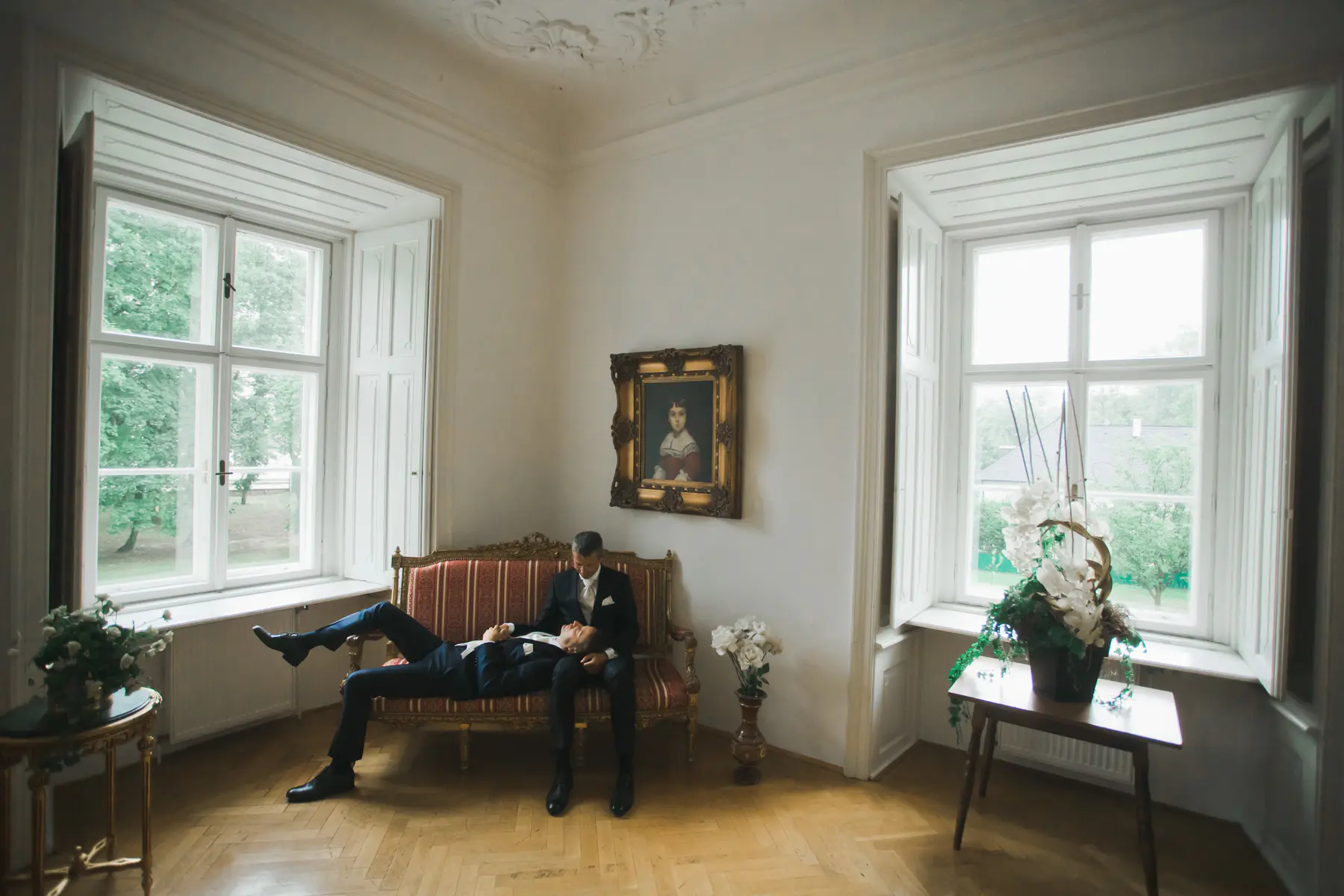 Two grooms in suits are sitting on a couch in a bright room before wedding. One man is lying down with his head in the other man's lap. There are two large windows in the room, one on the left and one on the right. A painting of a woman hangs on the wall between the windows.