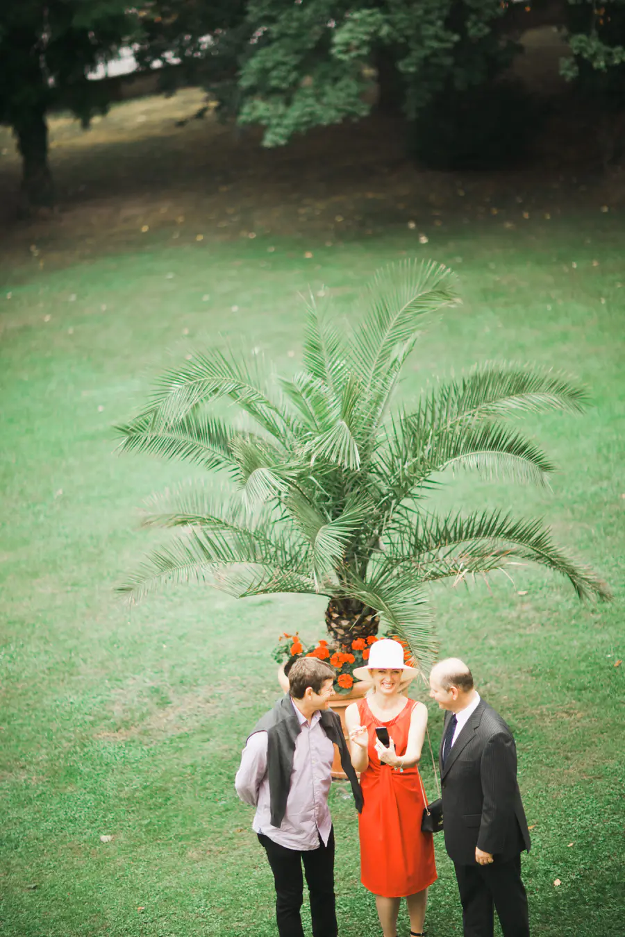 An overhead shot of two men and one woman standing on a grassy lawn. The woman, in a red dress and white hat, holds up a smartphone which all three are smiling and looking at. A potted palm tree with red flowers is behind them.