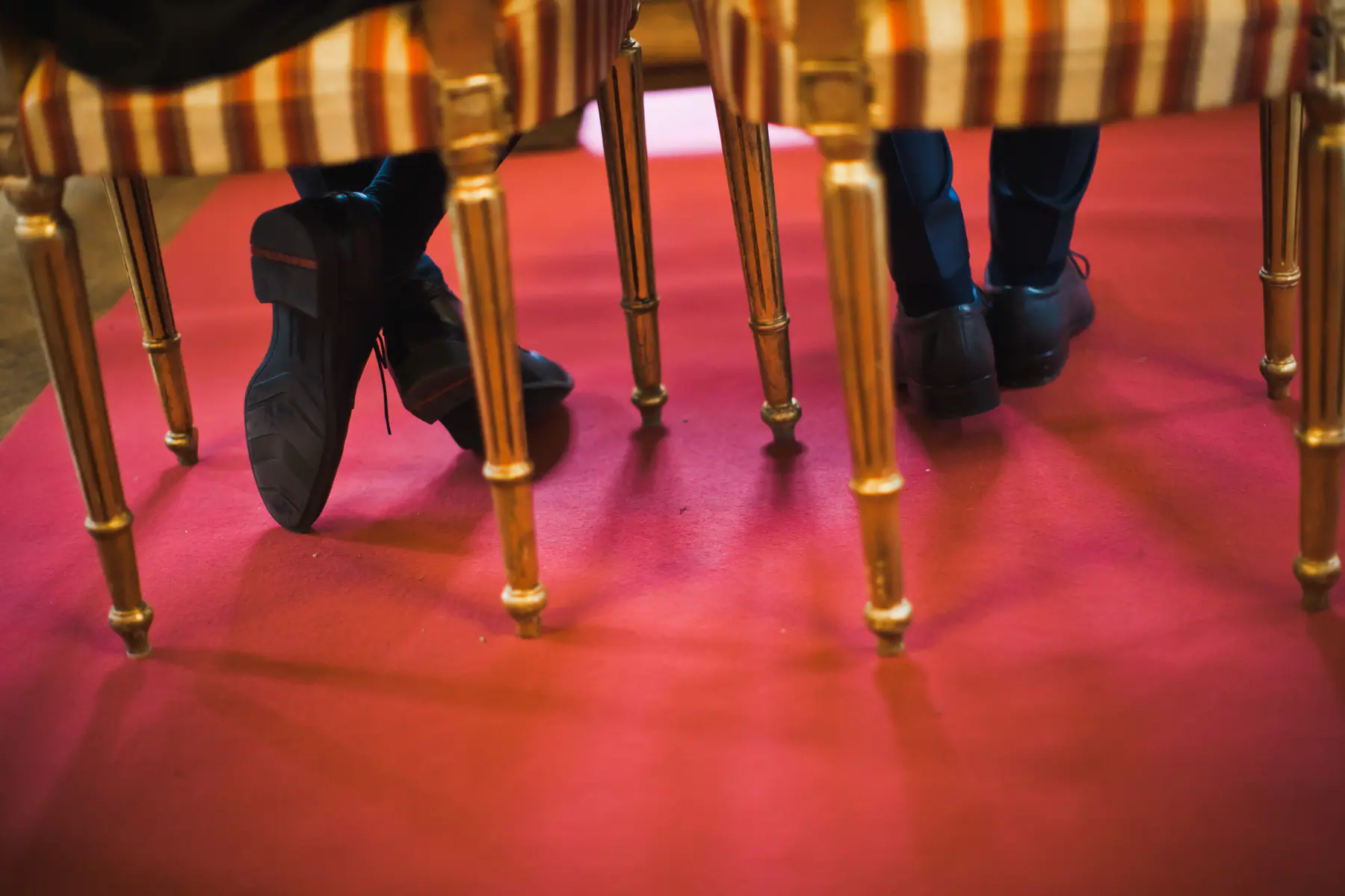 A low-angle view shows two people's feet in dark shoes on a red carpet, seen from under chairs with ornate golden legs. One person's feet are crossed, revealing the