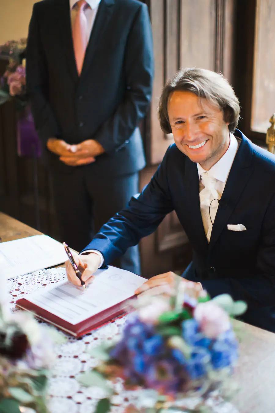 A man in a dark blue suit and white cravat signs an official document, smiling widely at the camera. Another man in a suit stands behind him, out of focus. 