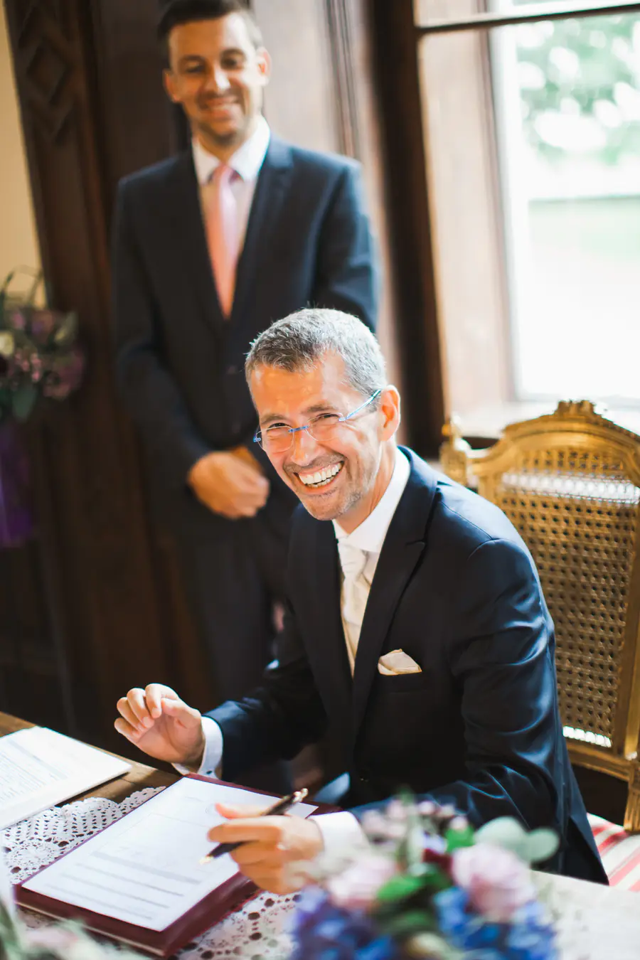 A man in a dark blue suit and white cravat signs an official document, smiling widely at the camera.