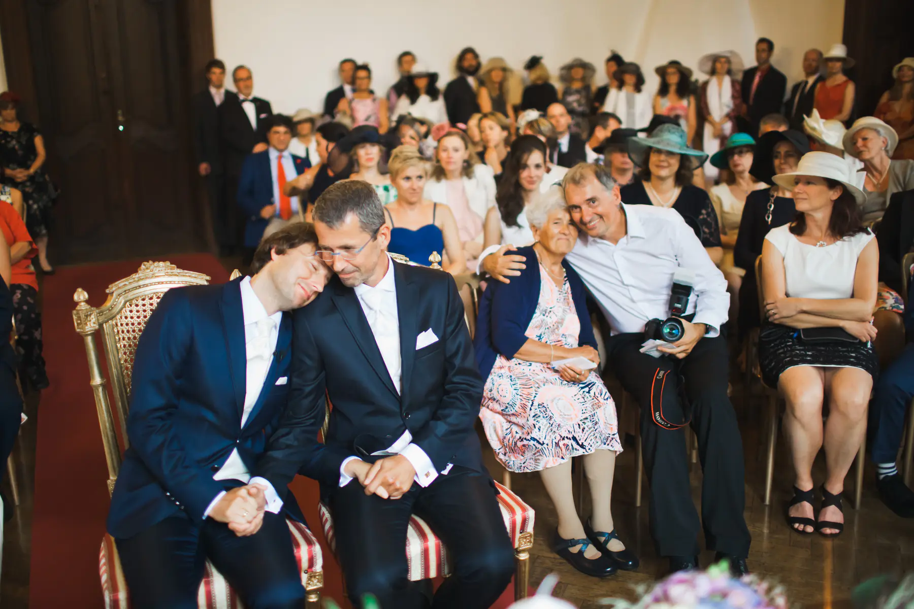 Two men in dark suits, likely grooms, sit intimately at what appears to be a wedding ceremony. One rests his head on the other's shoulder as they hold hands and smile softly
