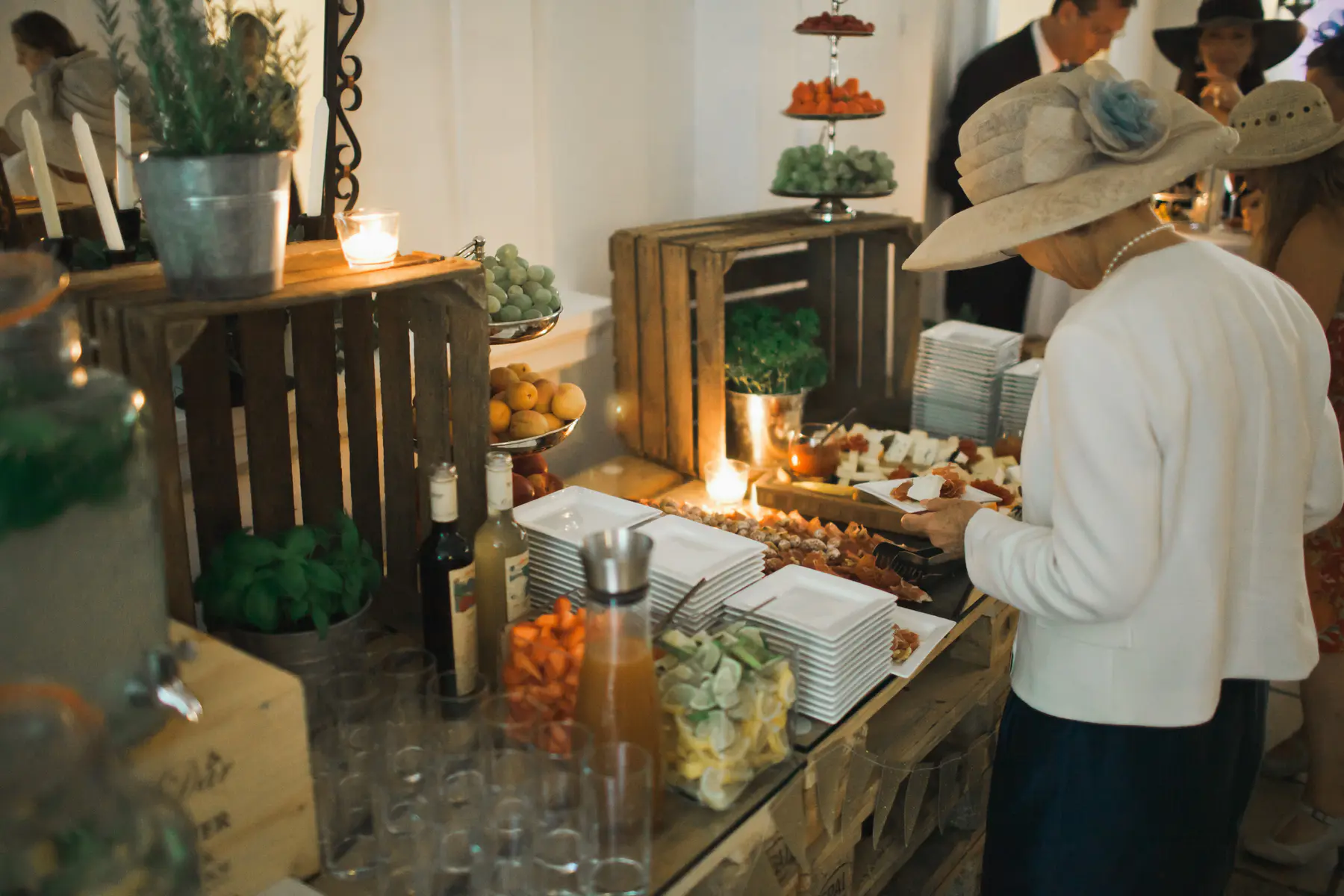 A woman in a large cream hat and white jacket serves herself from a rustic buffet table laden with a variety of finger foods, drinks, and stacks of plates. Decorative plants and candles adorn the table