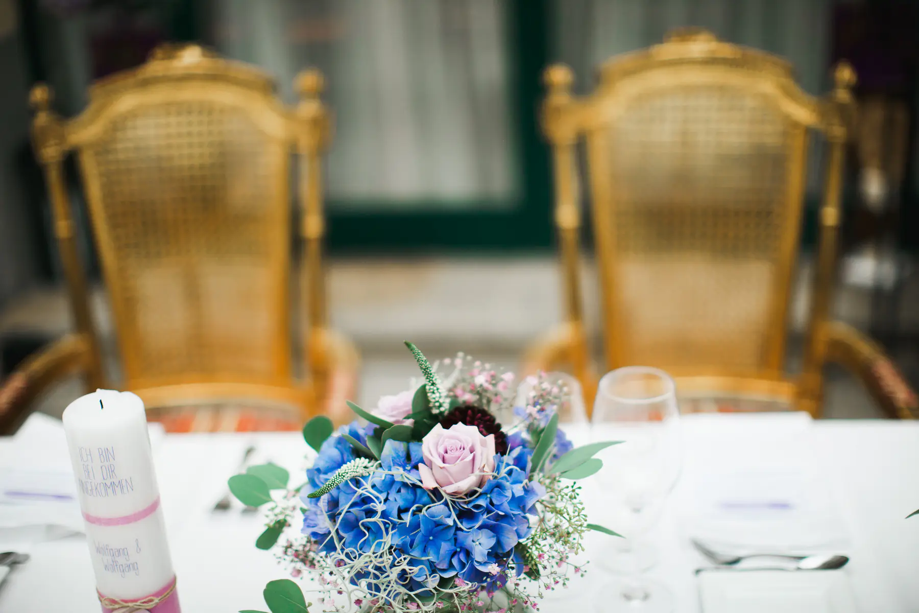A white candle with pink ribbon and text sits on a table next to a bouquet of blue hydrangeas and pink roses. Two ornate, golden chairs are blurred in the background.