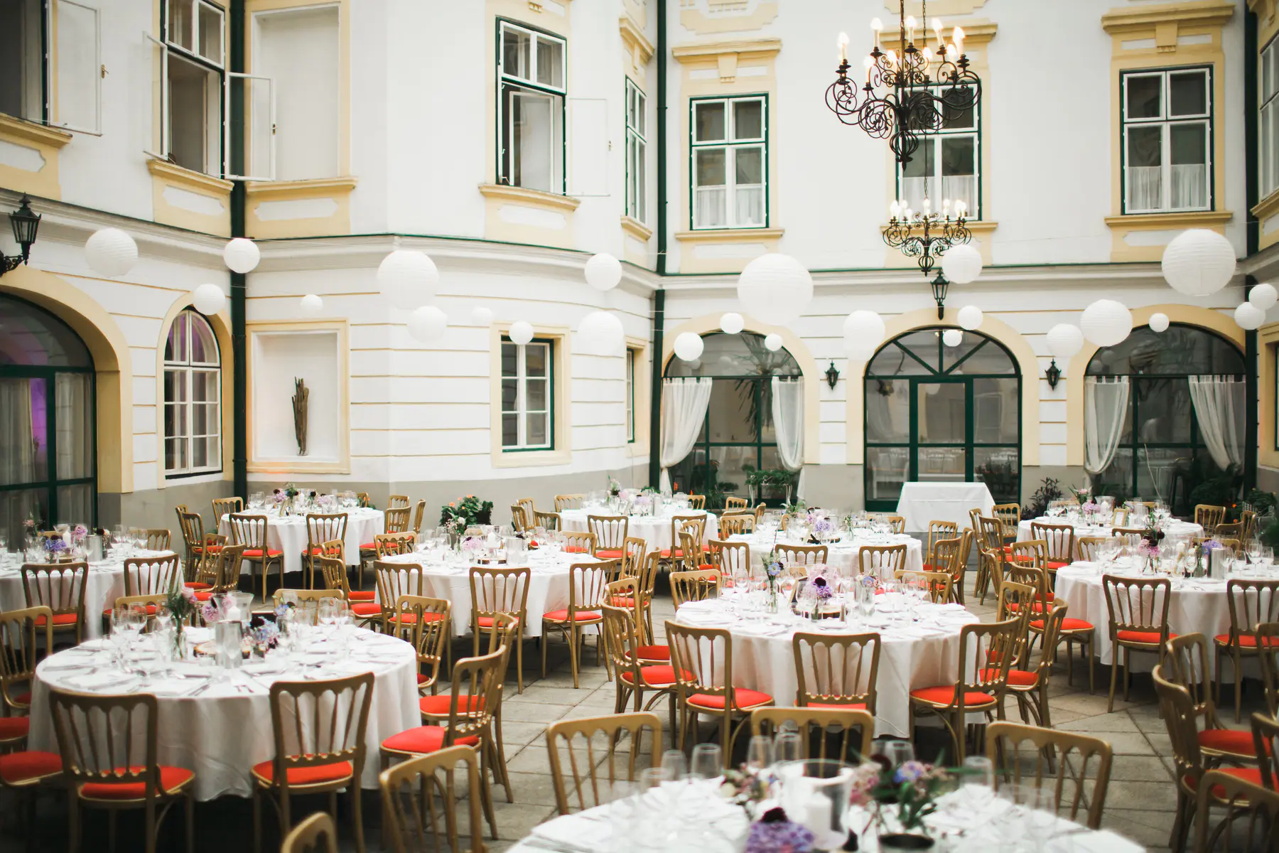 A formal outdoor dinner party is set up in a courtyard. Round tables draped in white cloths are surrounded by chairs with red cushions. White paper lanterns hang from the building's exterior, and elaborate chandeliers are suspended from the ceiling.
