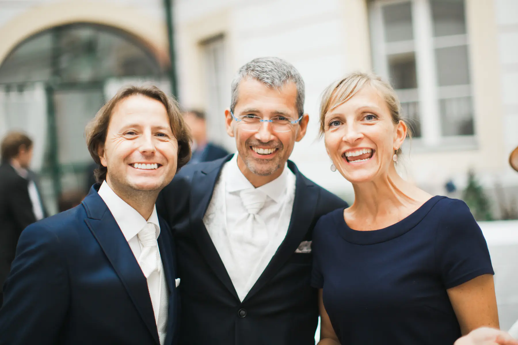 Three people, two grooms and a woman, smile at the camera. The man on the left wears a navy suit, white shirt, and white tie. The man in the center wears a dark suit, a patterned white vest, and a white tie, with blue-rimmed glasses. The woman on the right has blonde hair and wears a navy blue dress.