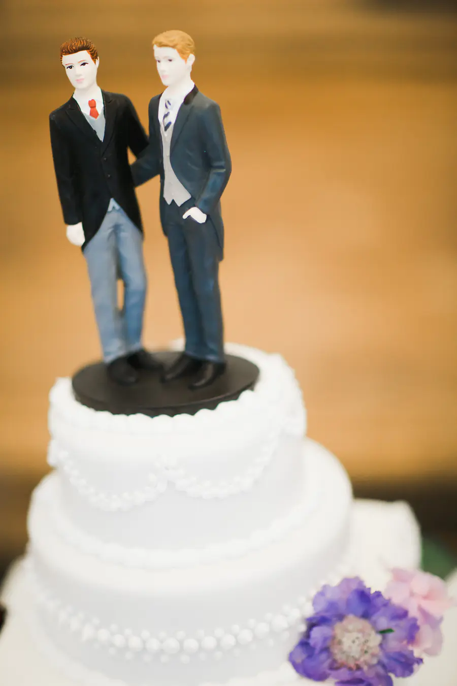 A multi-tiered white wedding cake topped with two male figures dressed in formal wear, signifying a gay wedding. The cake is decorated with white bead piping and purple and pink flowers.