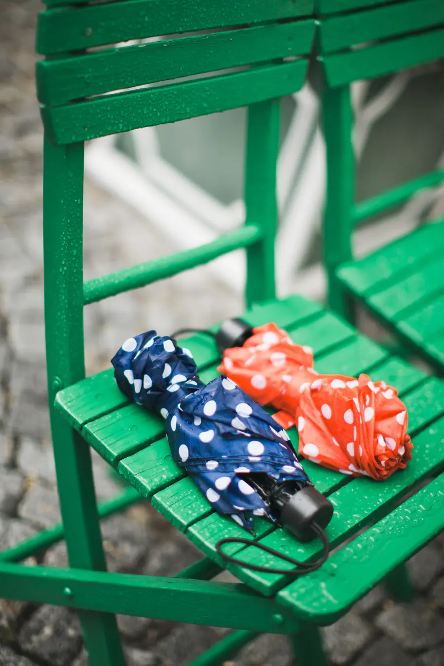 Two wet, folded umbrellas—one blue with white polka dots, and one orange with white polka dots—rest on a bright green wooden slatted chair covered in water droplets.