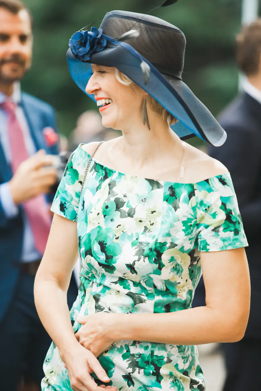 A woman in a blue wide-brimmed hat and a floral green and white off-the-shoulder dress laughs.