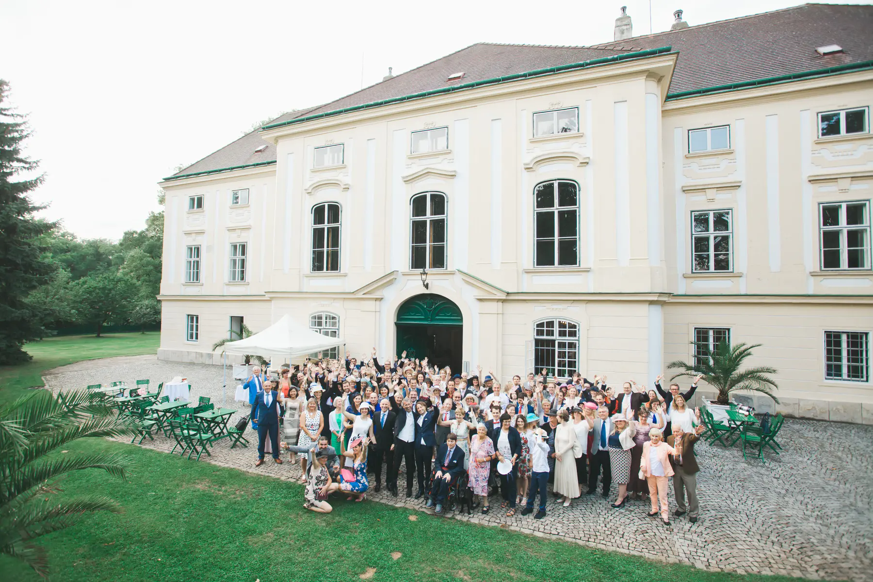 A large group of people, dressed in formal attire, stand in front of a pale yellow manor house. They are all looking towards the camera and many have their hands raised in a wave. The manor house has a green arched doorway in the center and many windows. There are trees in the background and a green lawn in the foreground.