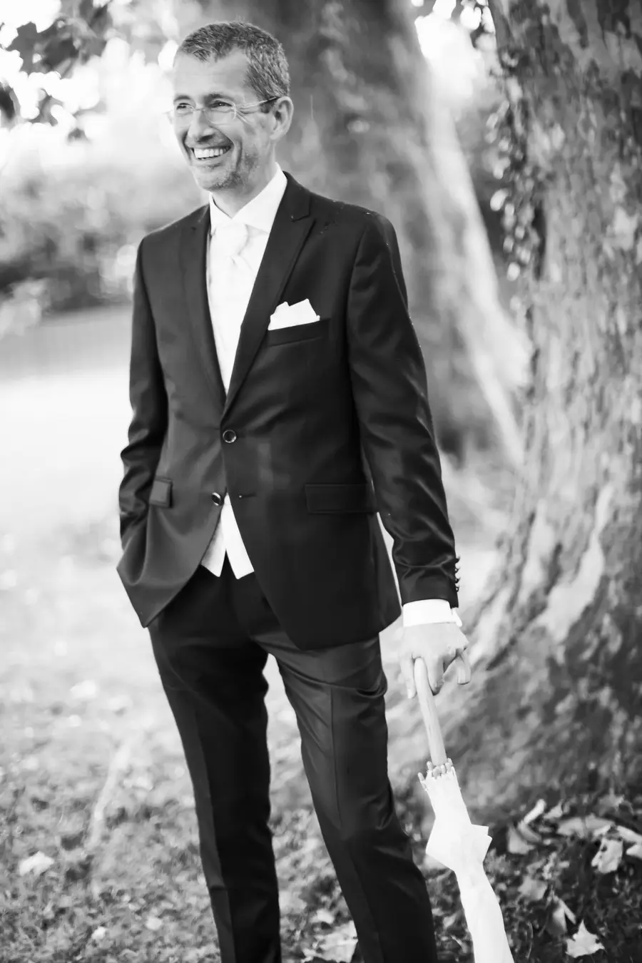 A smiling man in a dark suit, white shirt, and tie stands outdoors holding a white umbrella.