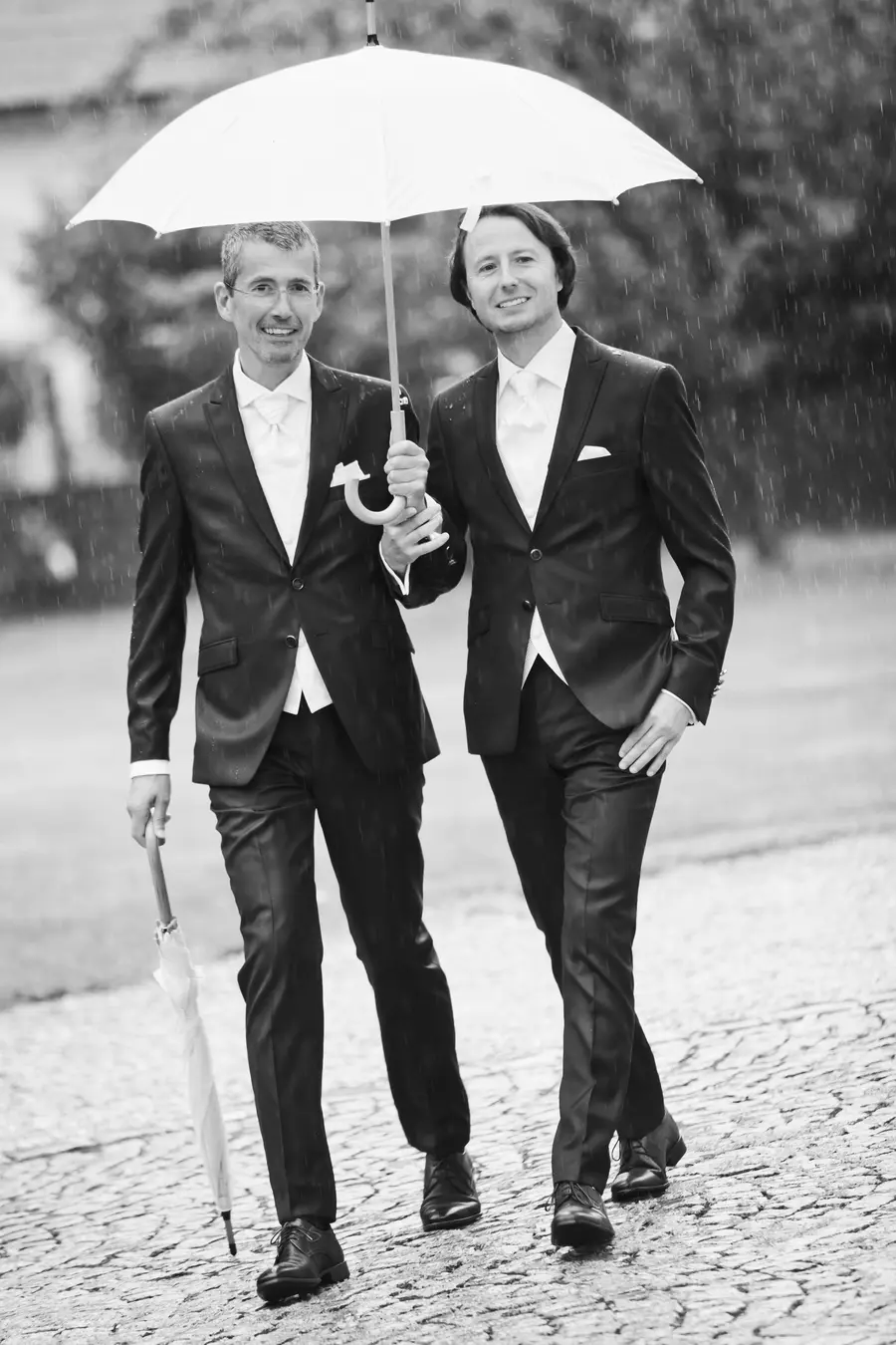 Two grooms in black suits and white shirts with ties walk under a white umbrella in the rain.Wedding photography Vienna
