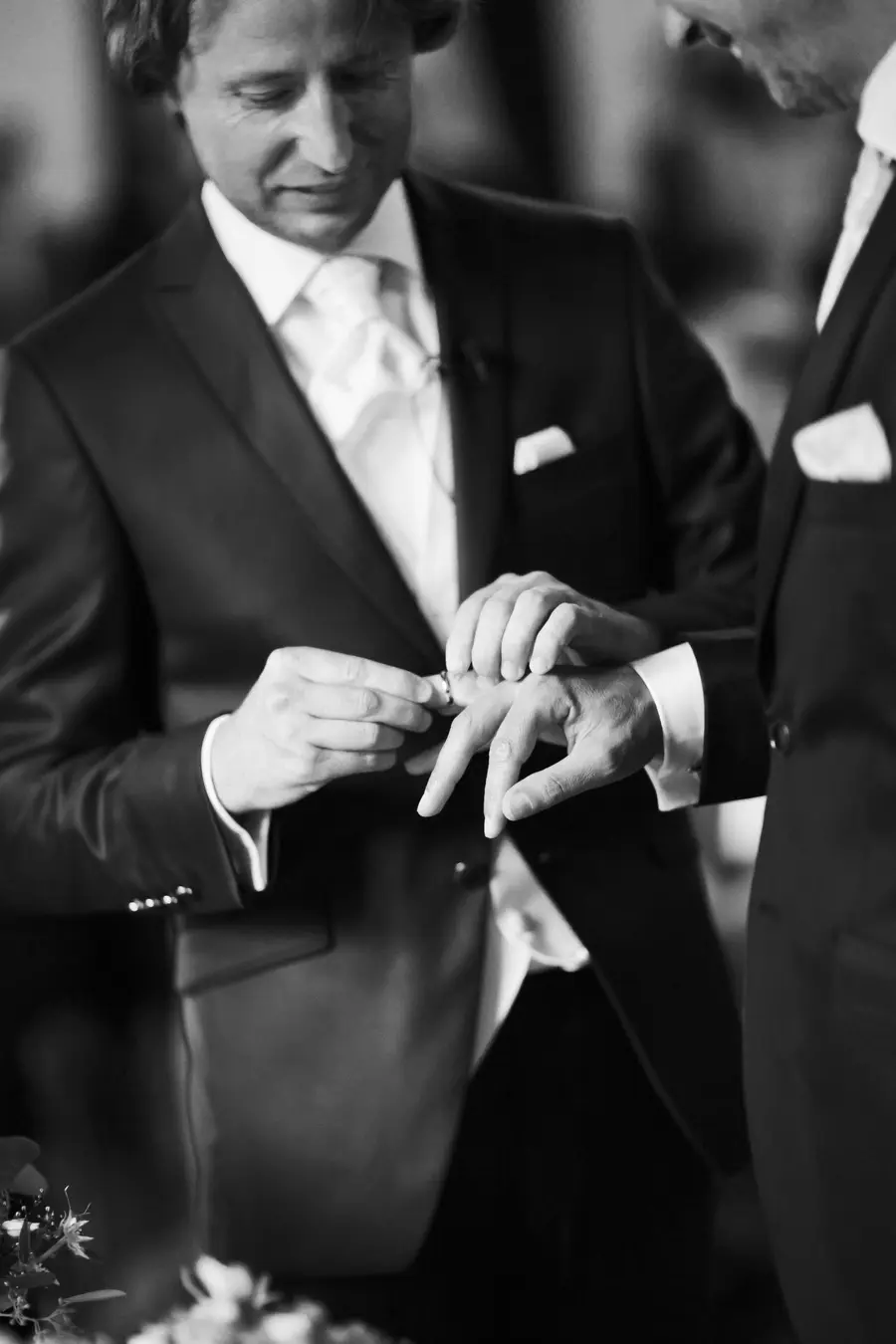 A black and white photo shows two men in dark suits. The man on the left is placing a ring on the finger of the man on the right, capturing a moment often seen during a