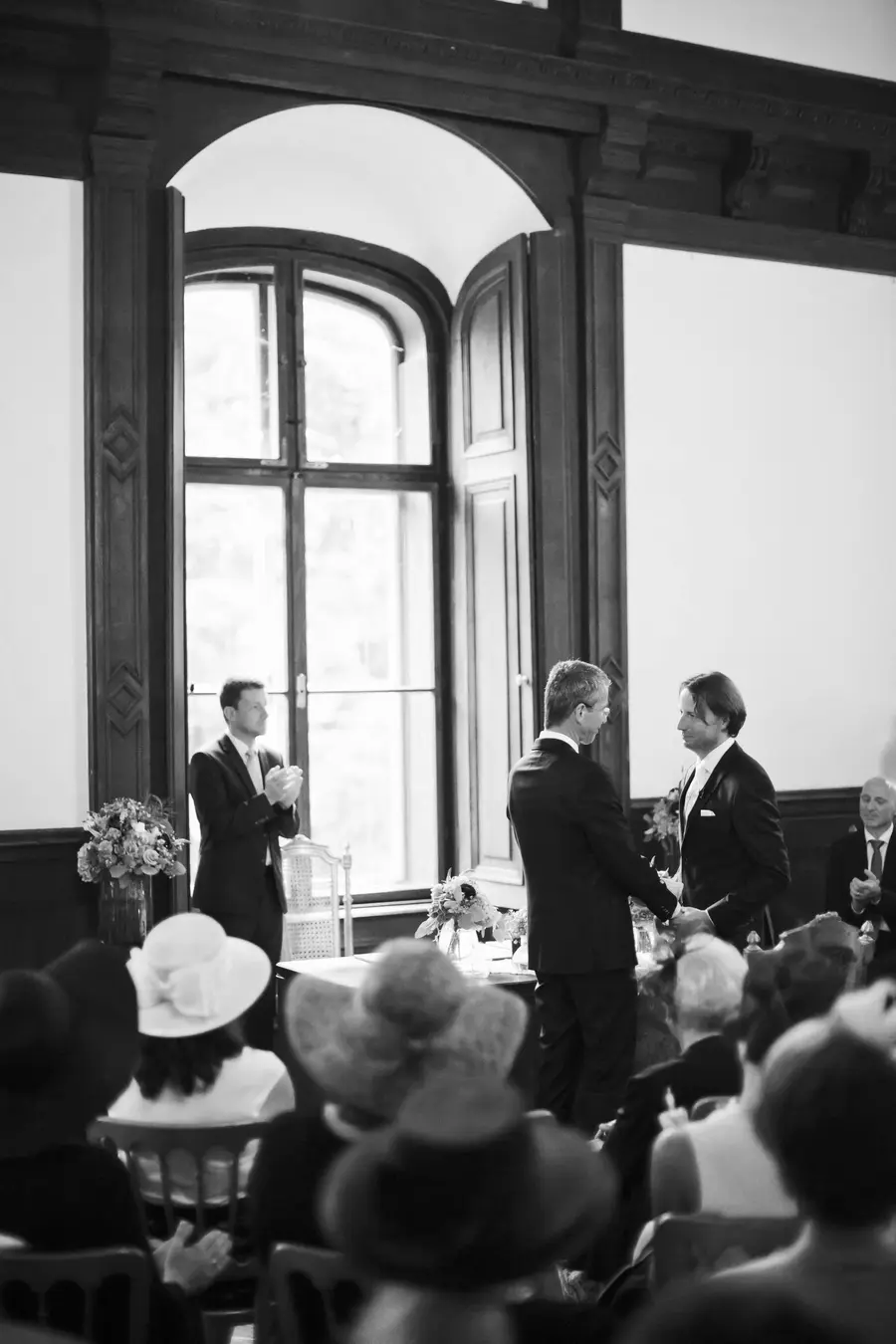 Black and white image of a formal ceremony. At the center, two men in suits face each other, holding hands. To the left, another man in a suit claps near a tall, arched