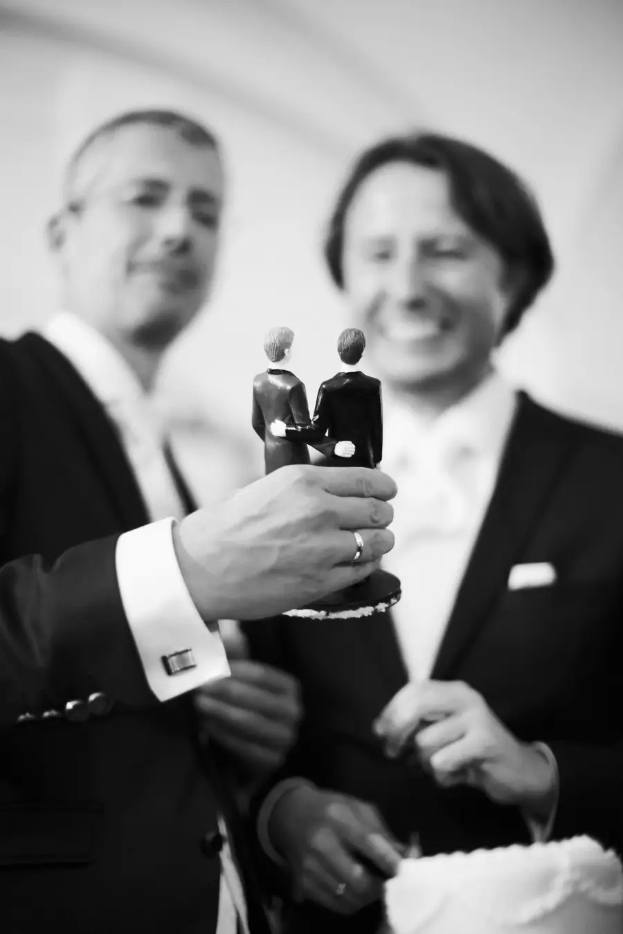 Black and white photo of a hand wearing a ring holding a wedding cake topper with two male figures arm-in-arm. Two blurred, smiling men in suits are in the background.