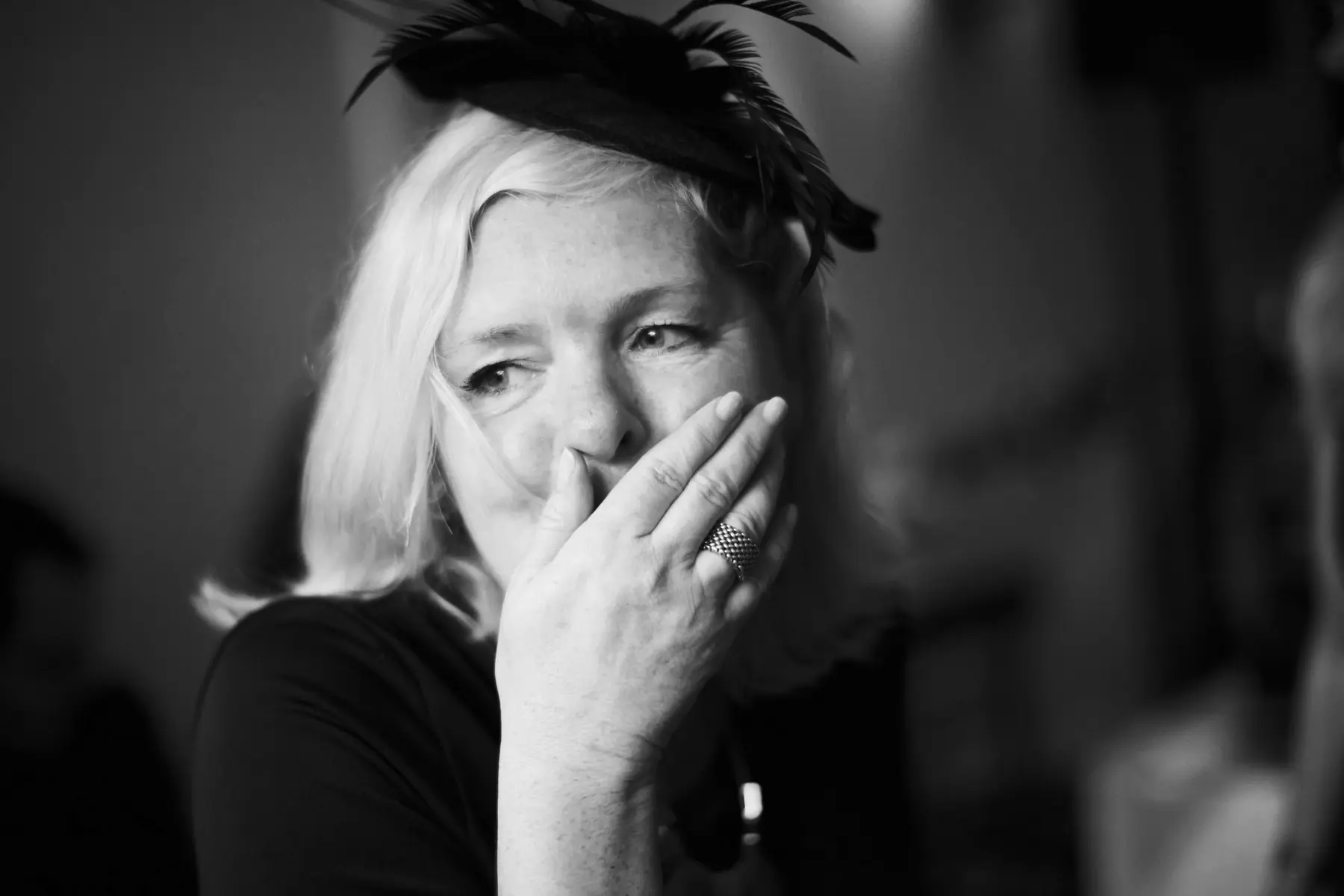 A candid close-up of a woman in an elegant wedding dress and hat, her hand covering her mouth in a moment of pure emotion.