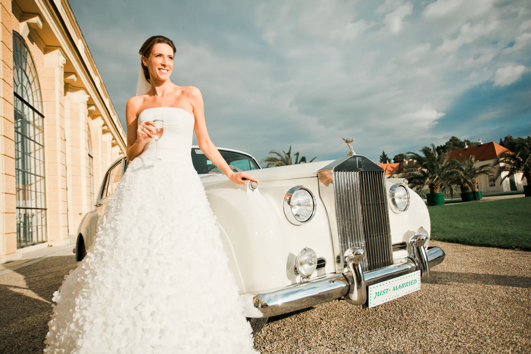 A smiling bride in a strapless white wedding gown holds a glass and leans on a classic white Rolls-Royce with a "Just Married" sign on its bumper. A grand building and palm trees are in the background.