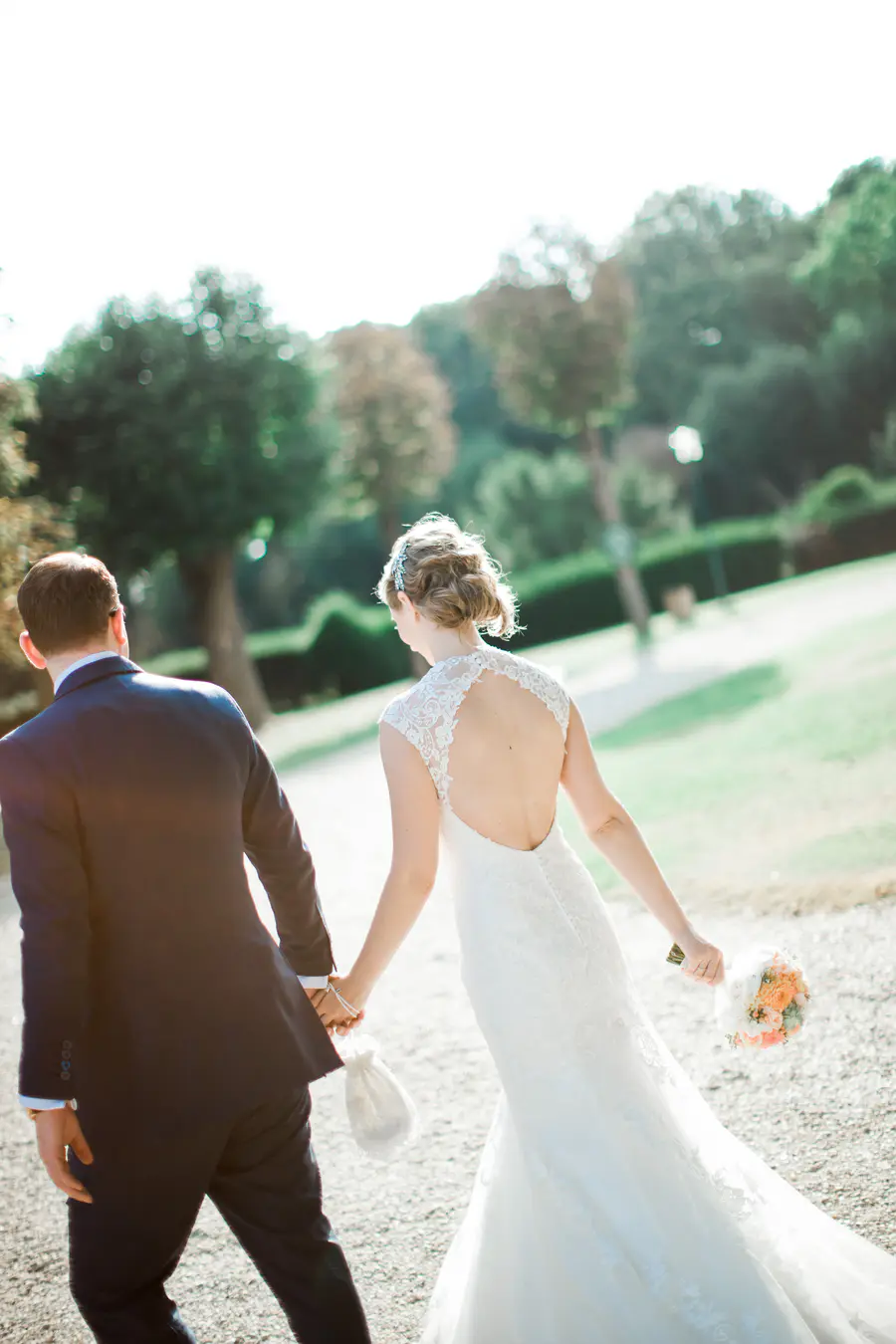 A bride and groom walk away from the camera, holding hands, with the bride in a white lace wedding dress and the groom in a dark suit.
