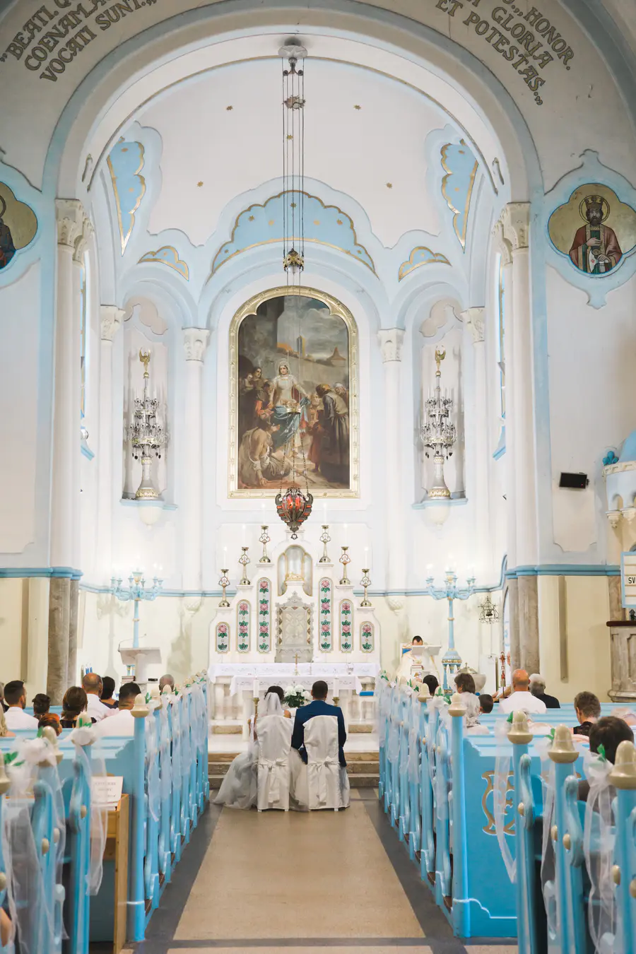 A wedding ceremony is underway in a bright, ornate church interior. A bride in a white gown and a groom in a dark suit are seated with their backs to the camera, facing the distant