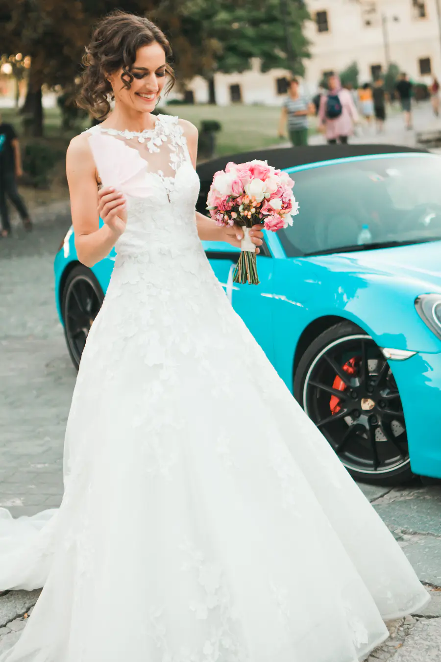 A smiling bride in a white lace wedding dress holds a pink and white bouquet and a pink card, standing next to a bright blue convertible Porsche outdoors.