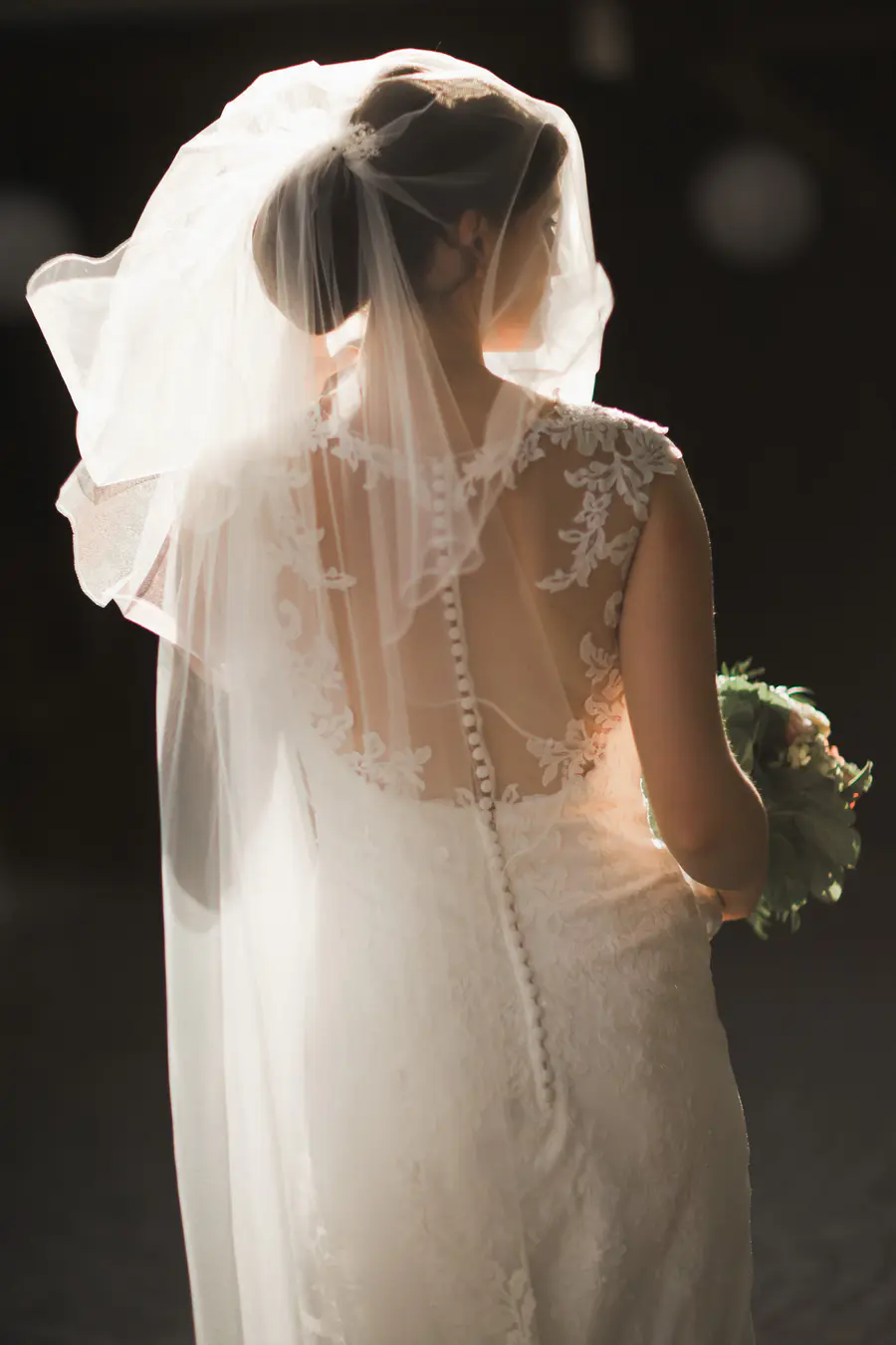 A bride seen from the back, wearing a white lace wedding dress and veil. Light atmosphere behing her