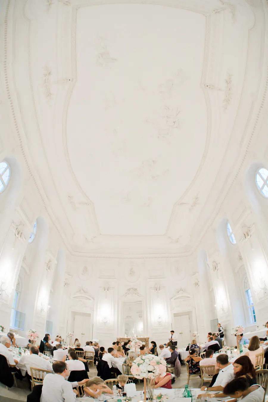 A wide-angle view of a formal event in a grand, ornate white room. Guests are seated at round tables adorned with floral centerpieces. The room features a high vaulted ceiling, intricate plasterwork on the walls, and several circular windows.