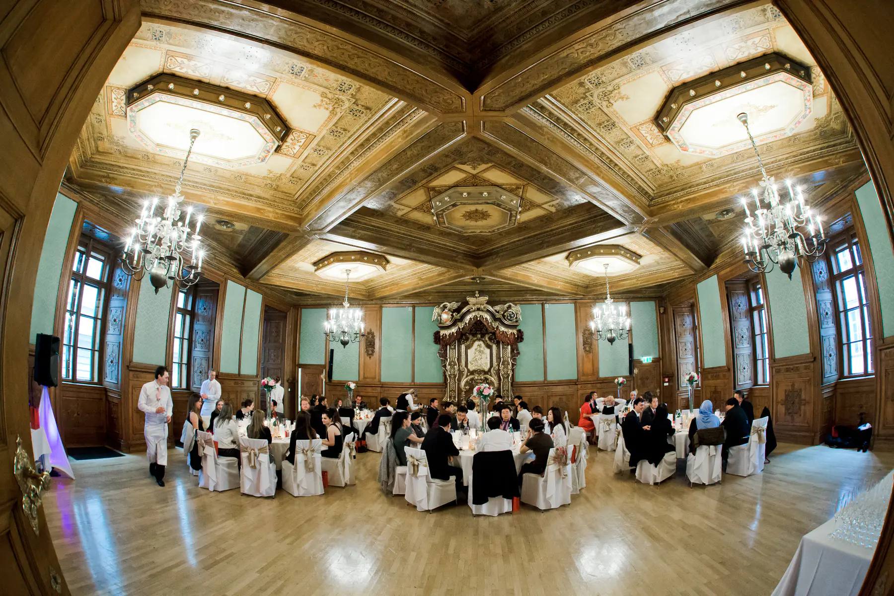 A grand, ornate ballroom with an intricately carved wooden ceiling and multiple chandeliers hosts a formal event. Guests are seated at numerous round tables with white tablecloths, while staff attend to them.