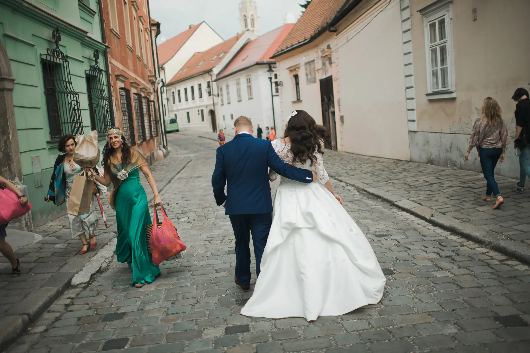 A bride and groom walk arm-in-arm down a cobblestone street lined with old buildings. The groom wears a dark blue suit, and the bride is in a white wedding dress. To their left, a woman in a green dress carries bags, and another woman in a patterned jacket walks beside her. In the background, other people walk further down the street.