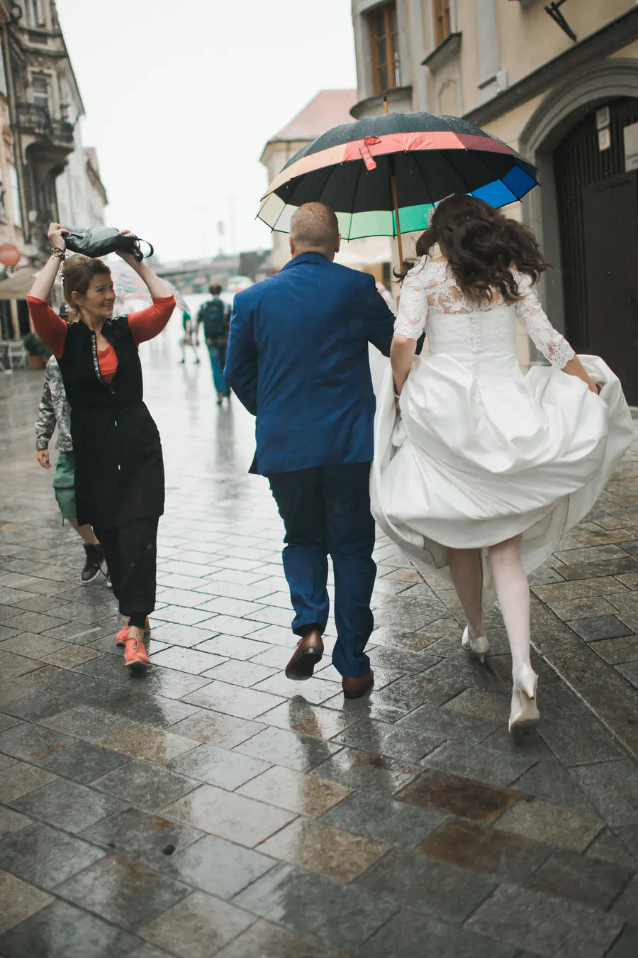 A bride and groom walk away from the camera under a colorful umbrella on a wet cobblestone street, with a woman in black holding a bag over her head in the foreground.