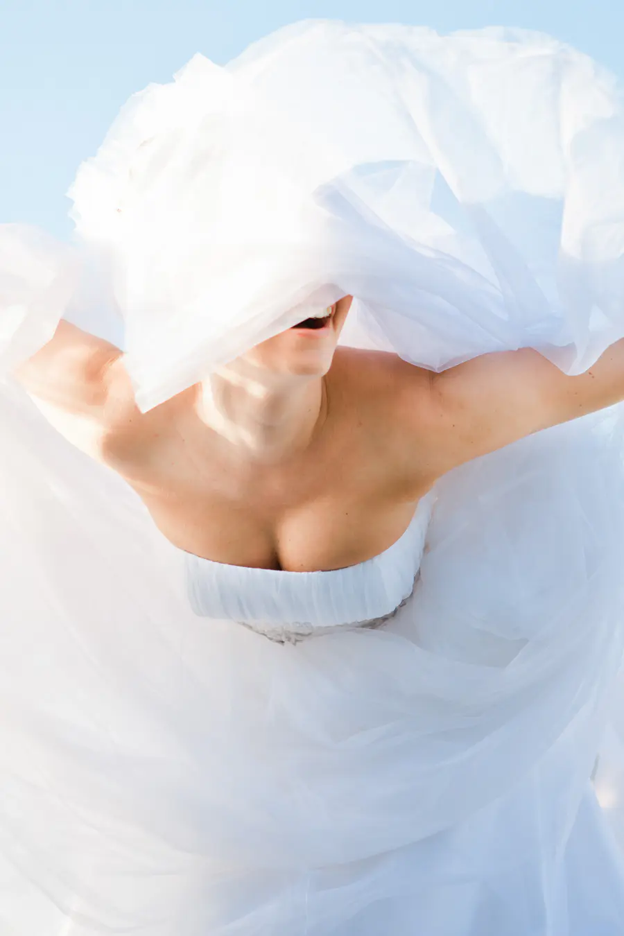 A woman in a white strapless wedding dress looks up, smiling widely, as voluminous sheer white fabric (likely a veil or part of her dress) billows around her head, partially obsc