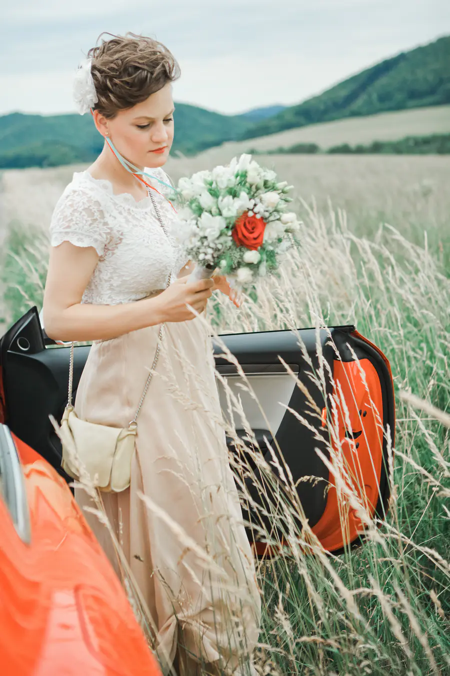 A woman in a white lace top and long beige skirt holds a white and red rose bouquet. She stands in a tall grassy field next to an open orange car door, with rolling green hills in the