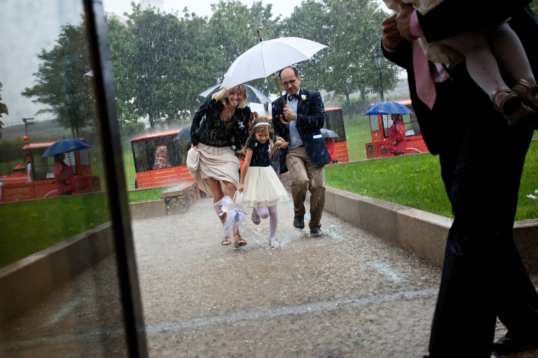 Authentic wedding photojournalism, candid emotions, no posing. A family runs in the rain under a white umbrella, holding hands.