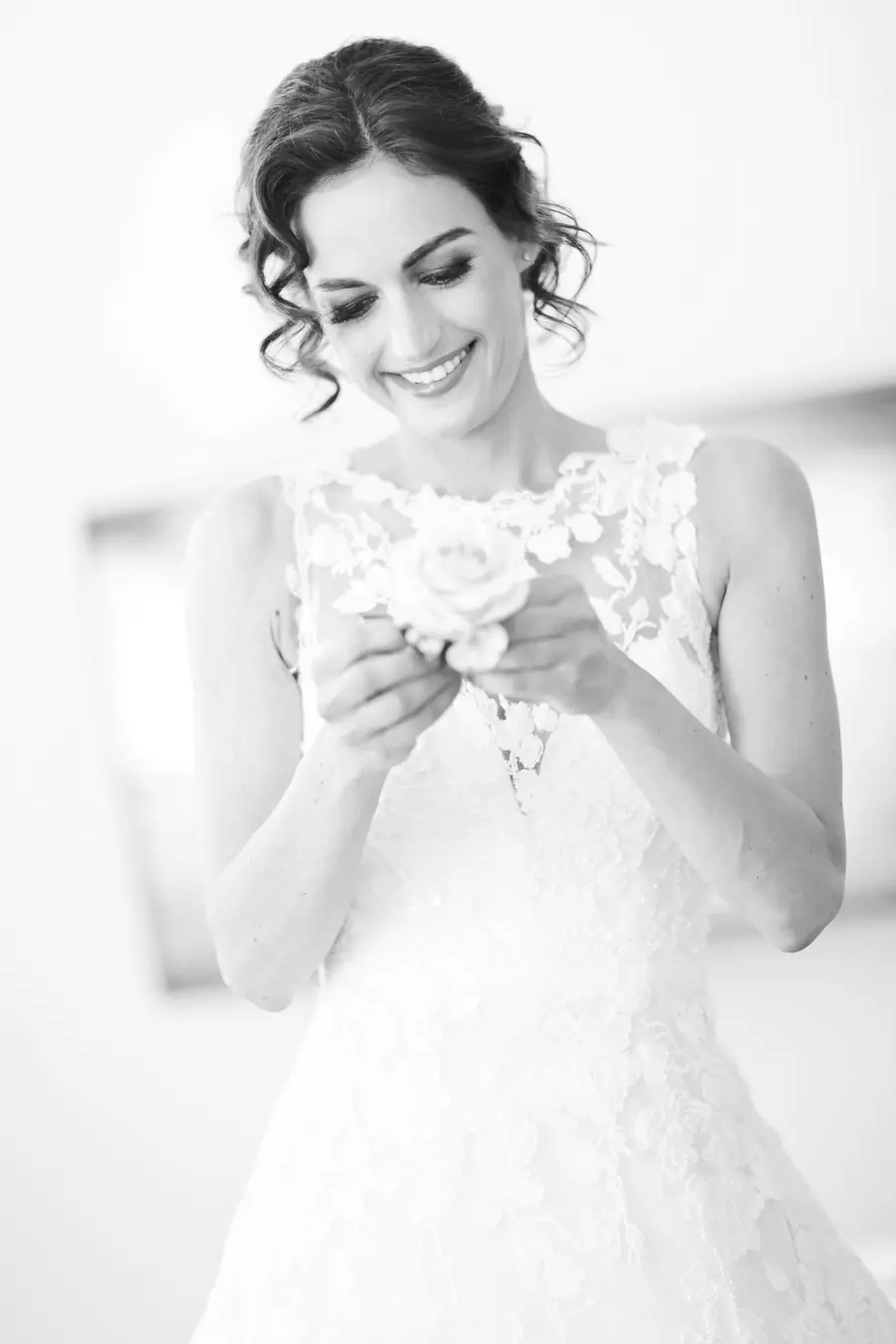 A black and white photo of a smiling bride in a lacy gown, looking down at a single rosebud held gently in her hands.